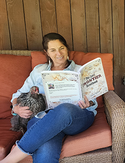 Suzanne sitting in an armchair holding a chicken and reading The Lost Frontier Handbook